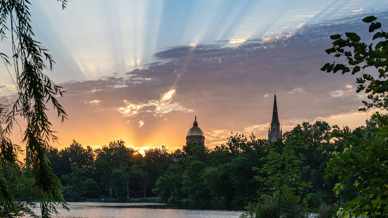 Sunrise over a lake with the Main Building and Basilica in the background. Sun rays are shining from behind low clouds.