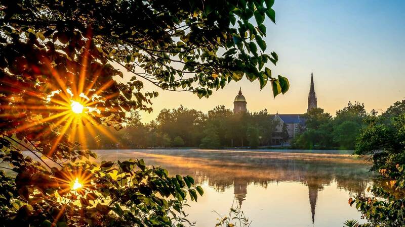 A view from across the lakes at Notre Dame. Rays from the sunrise appear behind the Main Building and Basilica which appear over the tops of the trees.