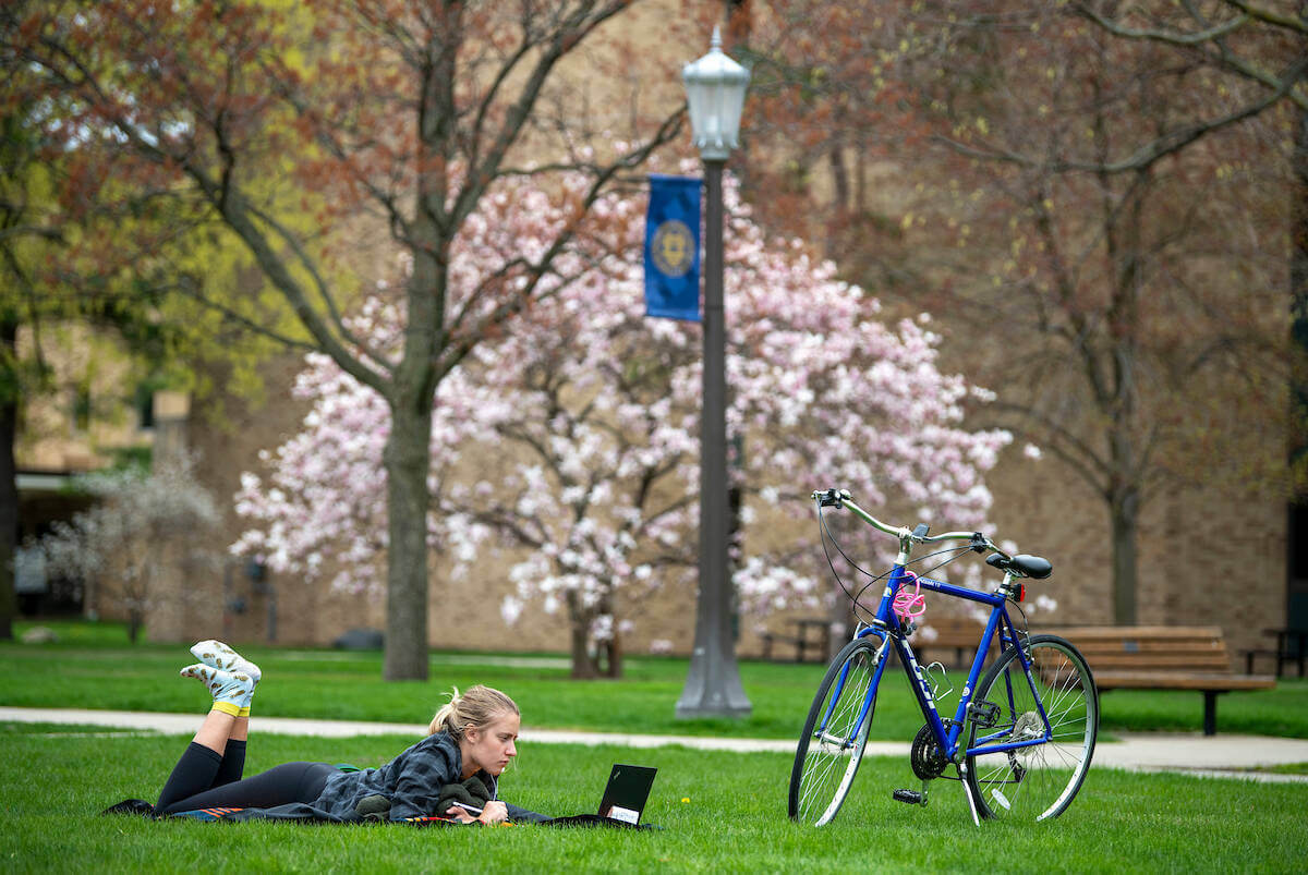 Student works on their laptop while laying in the grass by their bicycle